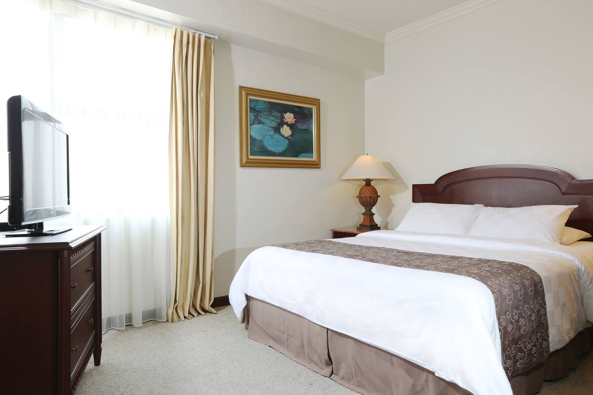A hotel room with a bed, a TV on a dresser, a painting on the wall, and a lamp on a nightstand.