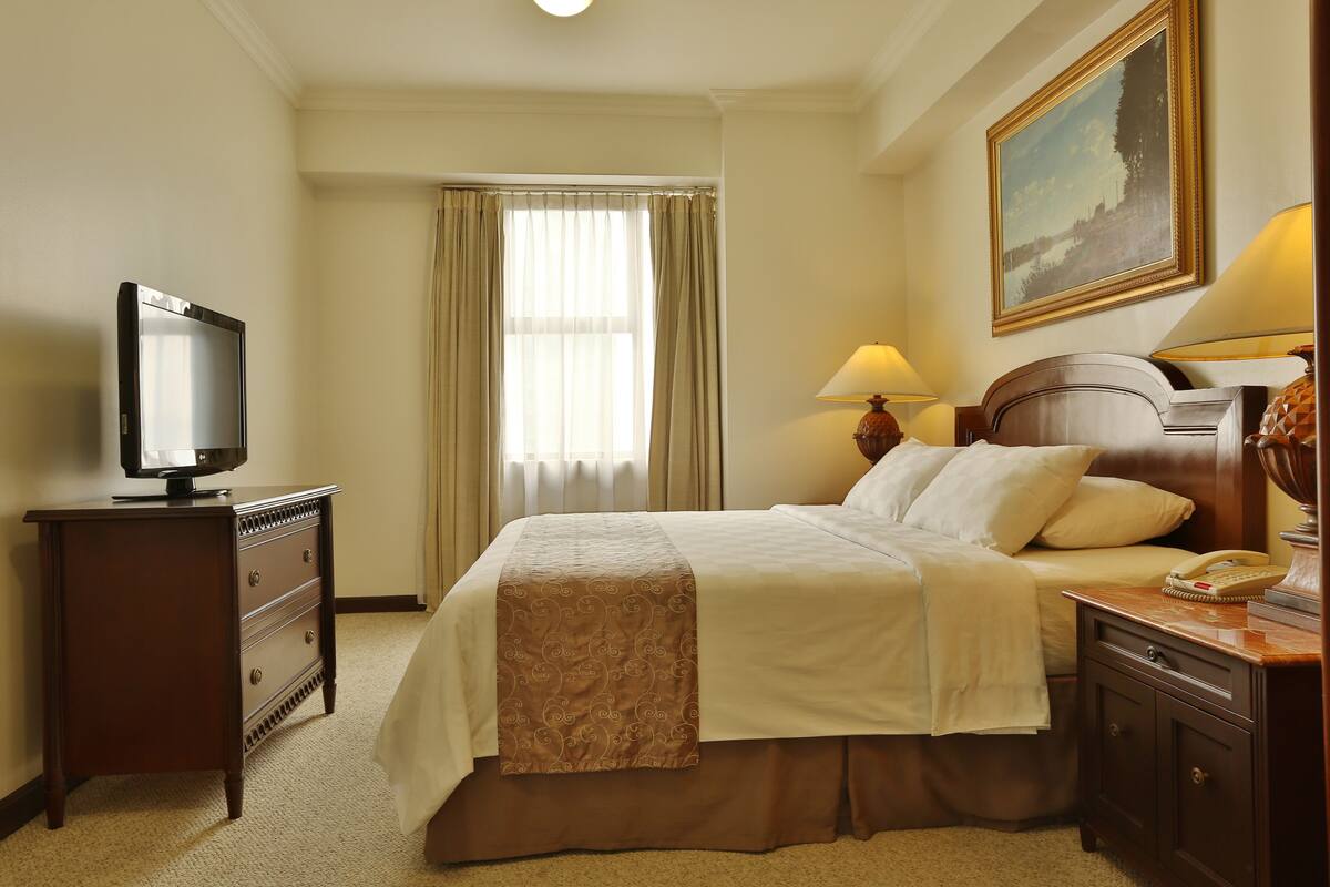 A hotel room with a bed, a TV, a desk with a lamp, and a painting on the wall.