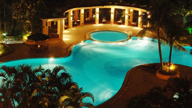 A nighttime view of a resort with a circular pool, a covered seating area, and palm trees. - Aryaduta Suites Semanggi (Jakarta)