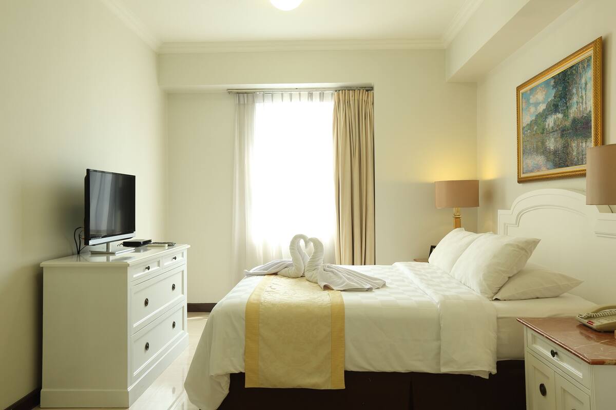 A hotel room with a bed, a TV on a dresser, a painting on the wall, and a window with curtains.