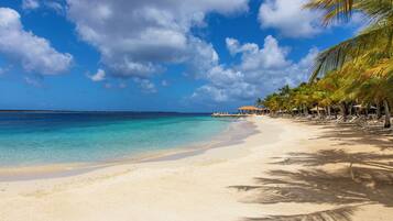 A beach with clear turquoise water, palm trees, and a distant building with an orange roof.
