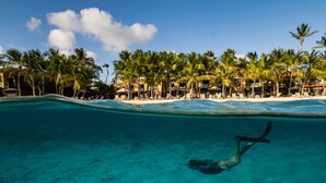 Visão subaquática de uma piscina turquesa límpida com um mergulhador, cercado por palmeiras e um resort na praia ao fundo.