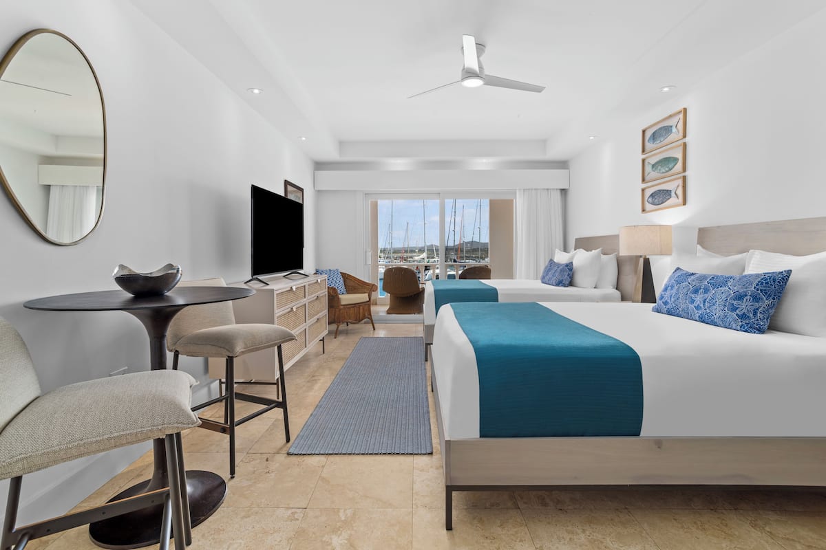 A hotel room with two beds, a desk, a chair, a TV, and a view of the marina.