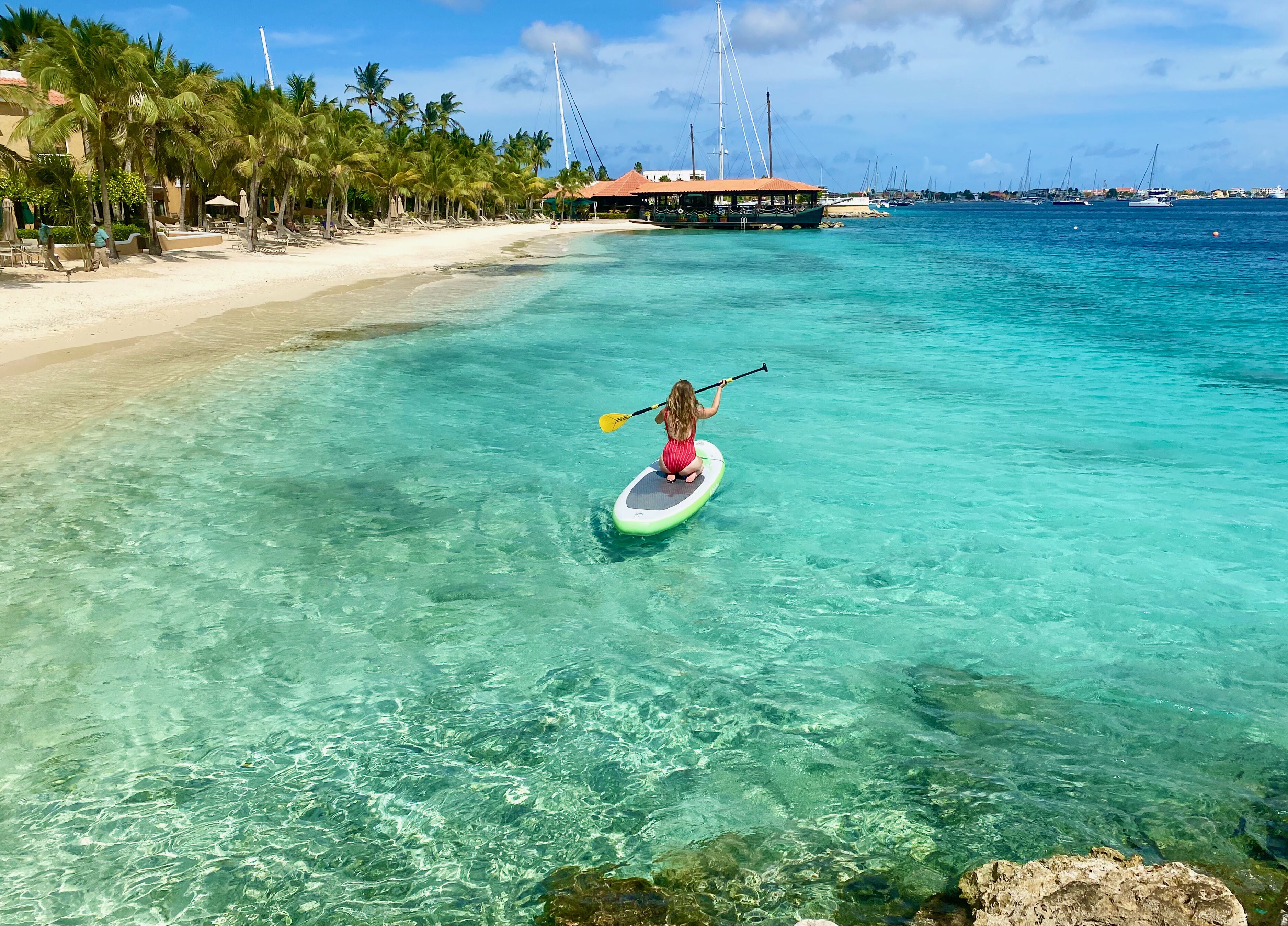 A beach with clear turquoise water, a paddleboarder, and a distant sailboat.
