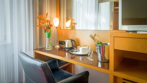 Ett hotellrum med ett skrivbord i trä, en stol, en tv, en vas med blommor, en vattenkokare, en kopp och ett glas.