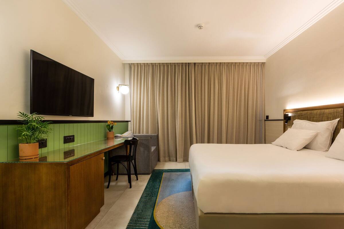 A hotel room with a bed, a desk with a chair, a television, and a window with curtains.