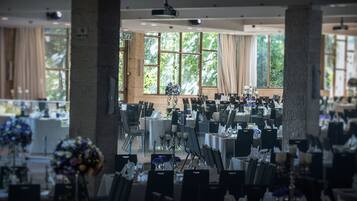 Un amplio salón de banquetes con varias mesas dispuestas para comer, grandes ventanales que dejan entrar luz natural y una estructura de pilares de piedra.