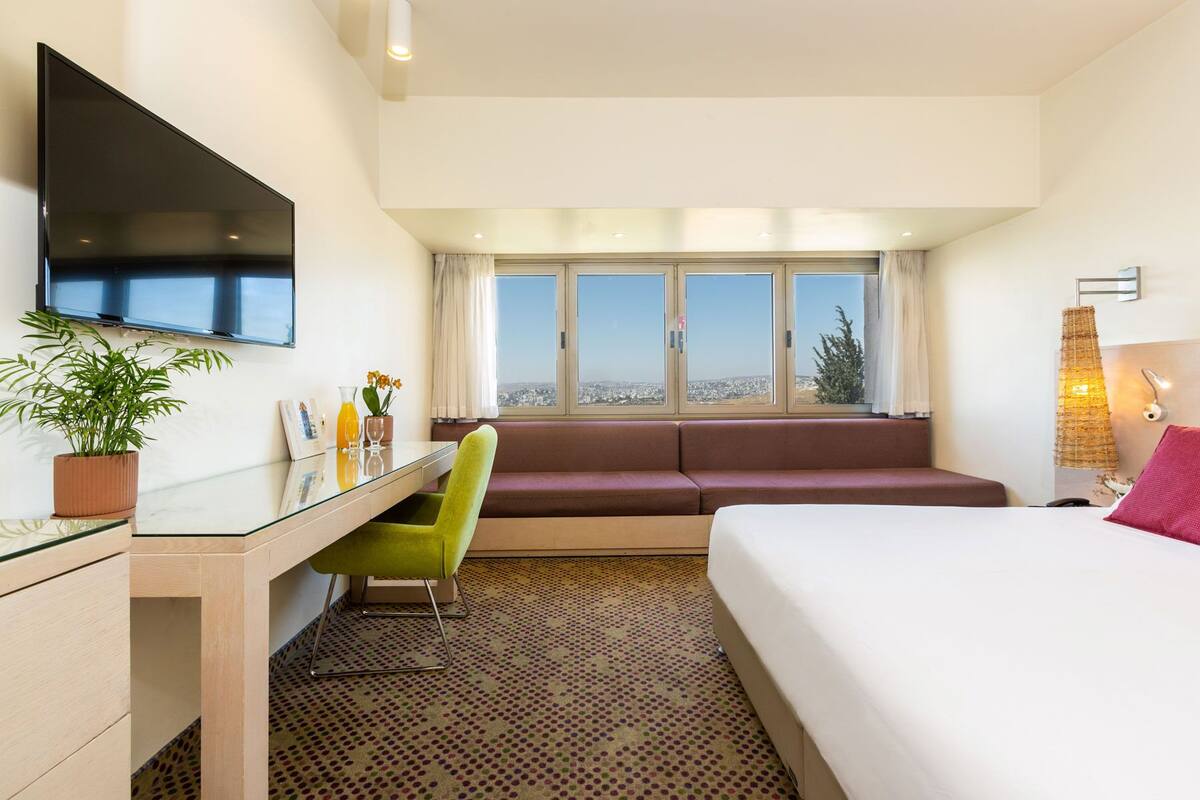A hotel room with a bed, a desk with a chair, a television, a window with a view, and a sofa.