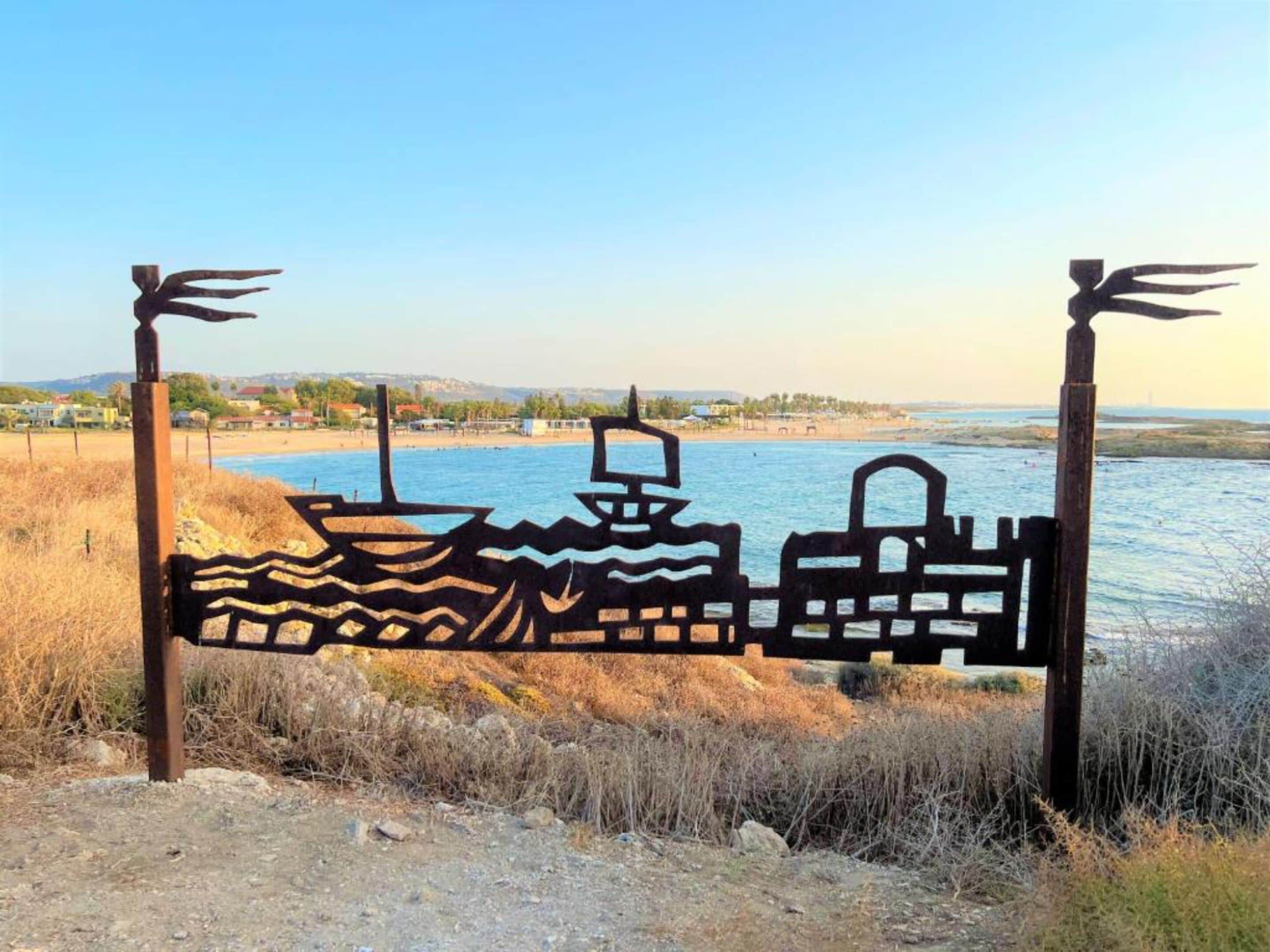 A metal gate with a wave pattern and a boat silhouette, set against a coastal backdrop with a clear sky.