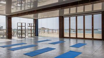 A spacious room with large windows overlooking a beach, featuring blue mats for yoga or stretching.