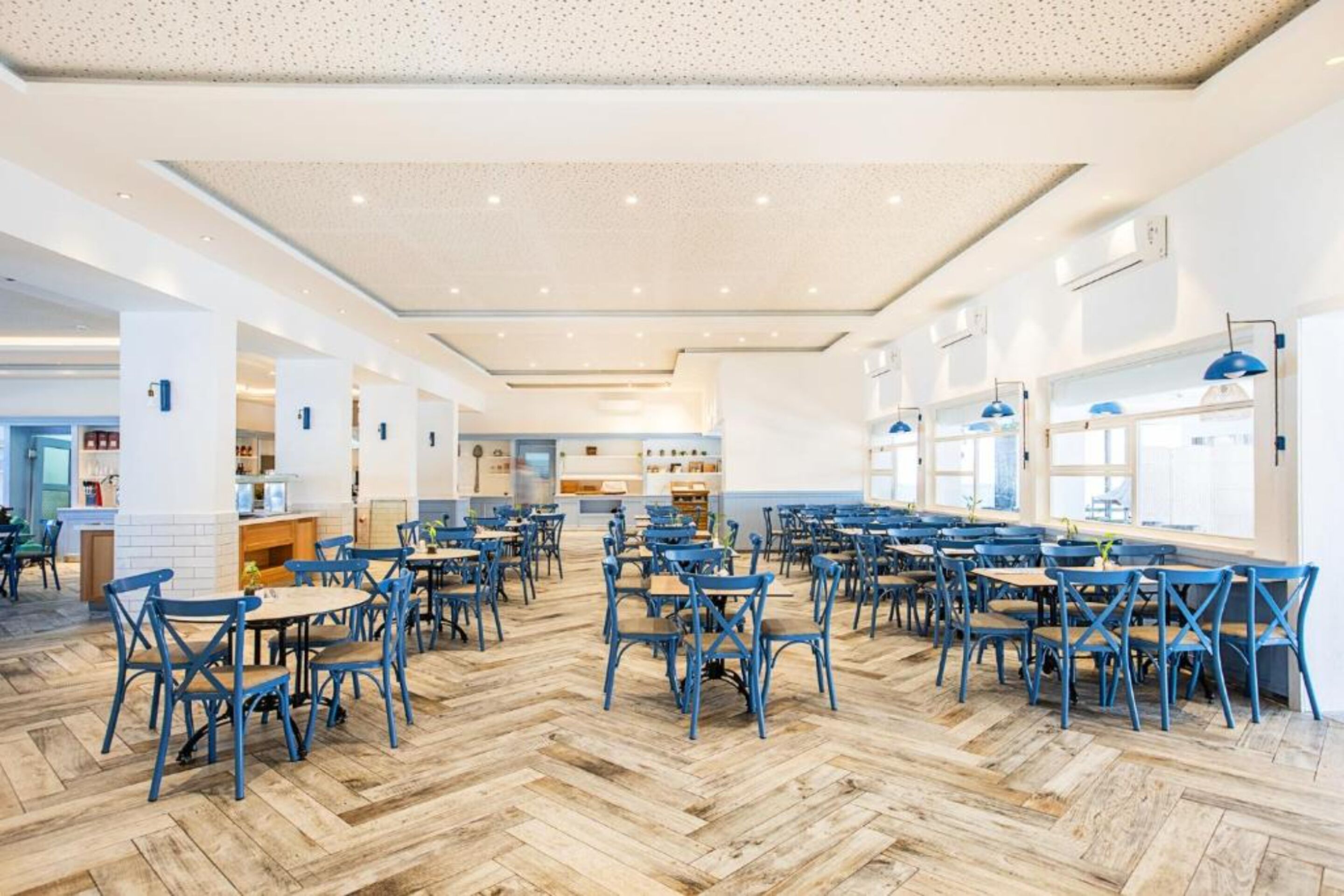 A spacious dining area with multiple tables and chairs, a herringbone patterned floor, and a modern ceiling design.
