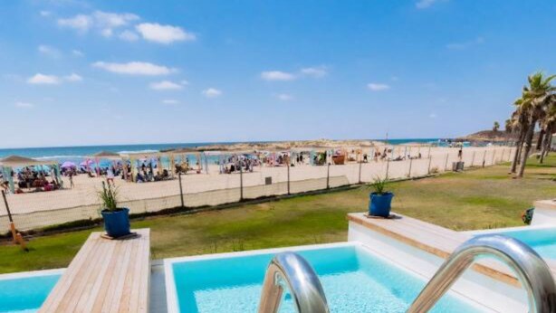 Una piscina con cubierta de madera y barandillas de metal, rodeada de una zona de playa con gente y palmeras.