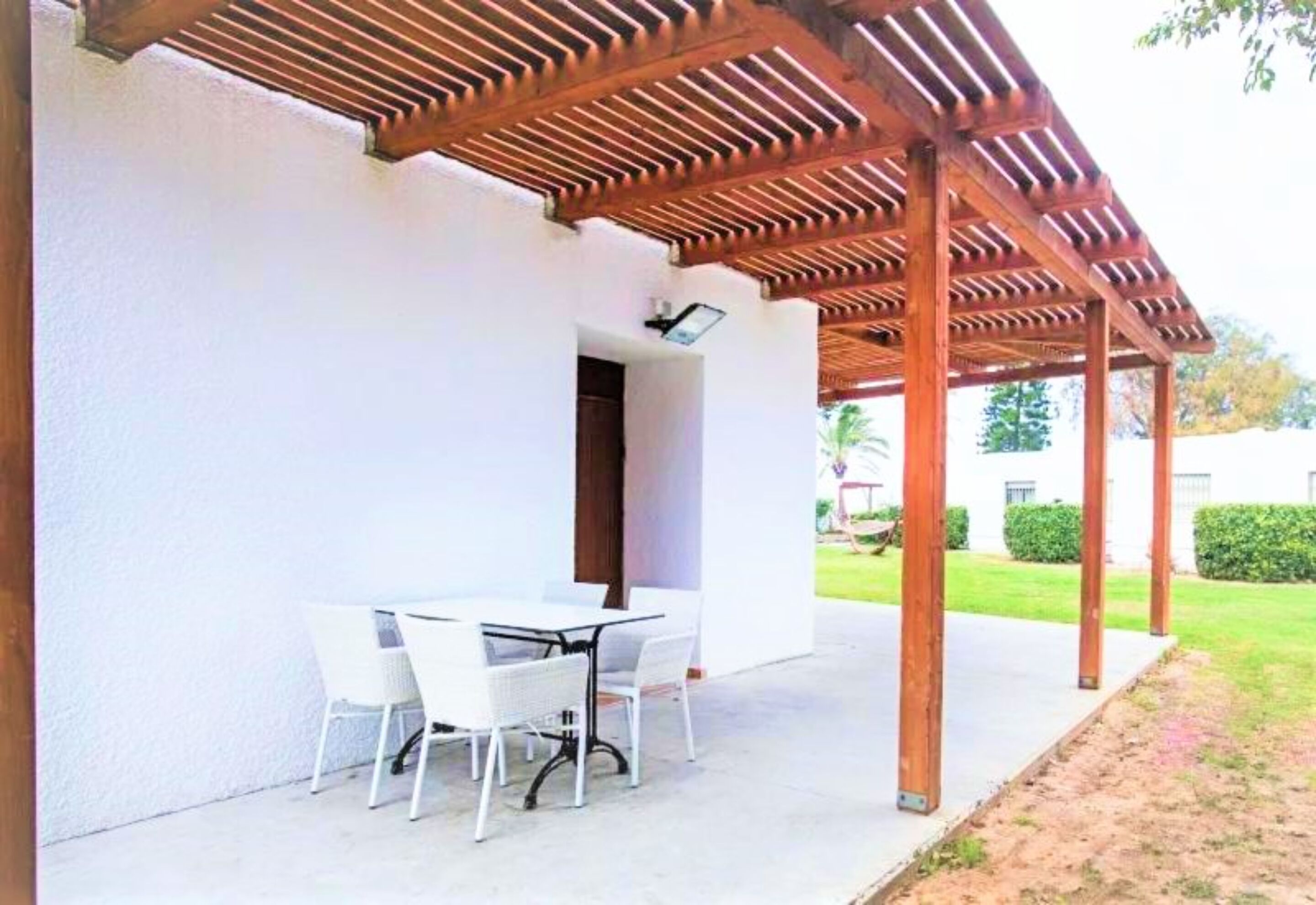 Een terras buiten met een houten pergola, een witte tafel en vier witte stoelen.