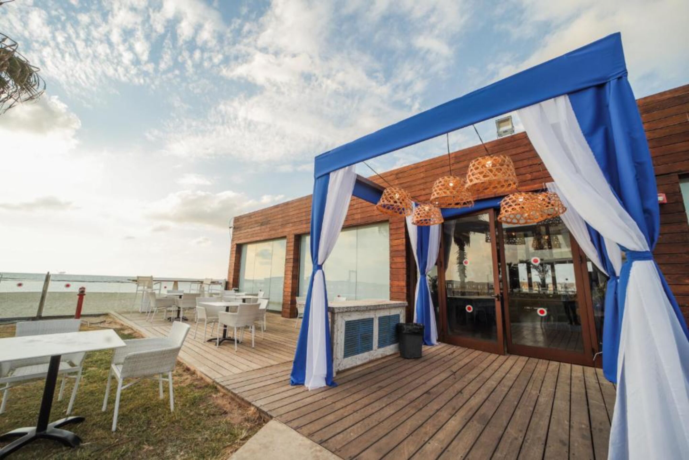 An outdoor patio area with a wooden deck, white and blue curtains, and hanging wicker lamps.