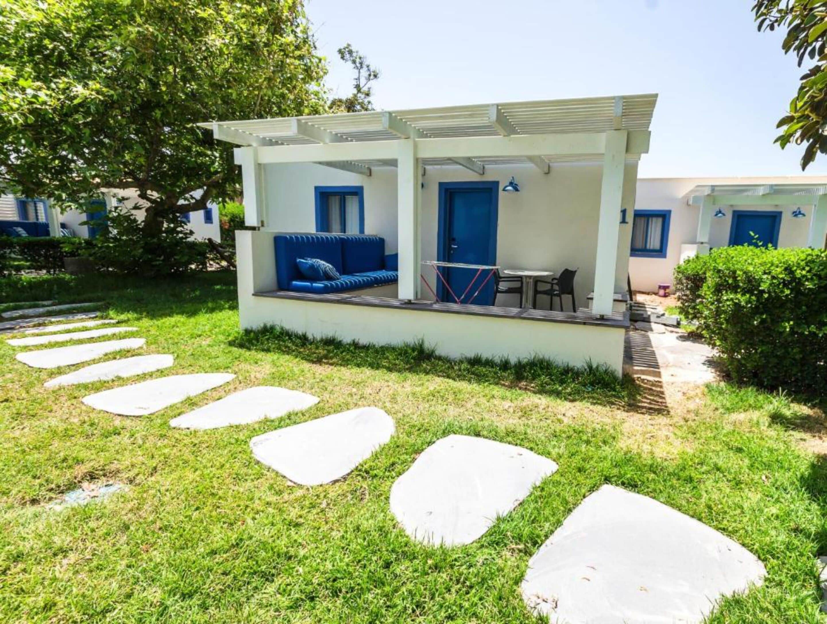Un área de estar al aire libre con techo, que incluye un sofá azul y una mesa, rodeada por una pérgola blanca y un camino de piedra.