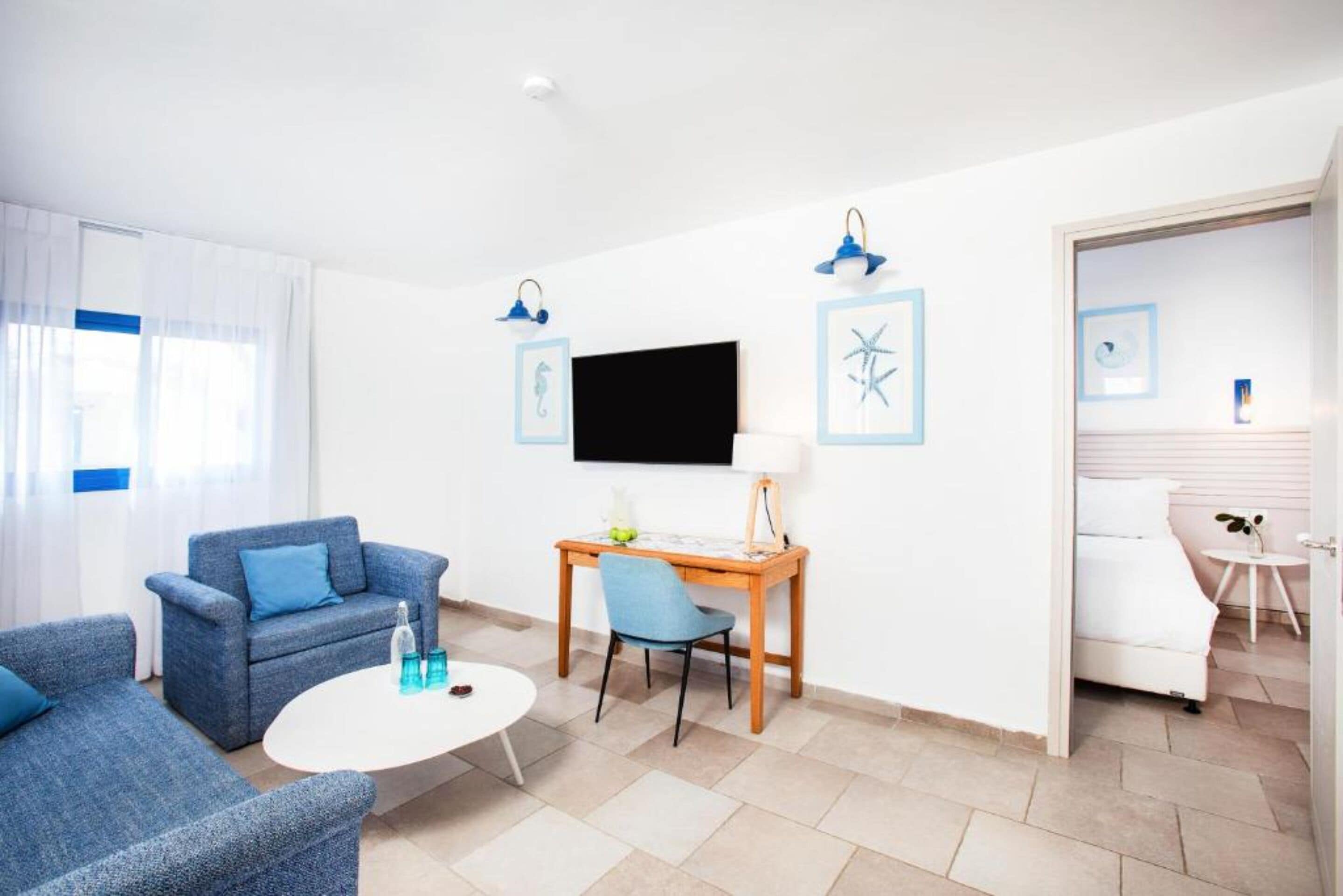 A modern hotel room with a blue sofa, a small round table, a desk with a chair, a TV mounted on the wall, and a bed with a white headboard.
