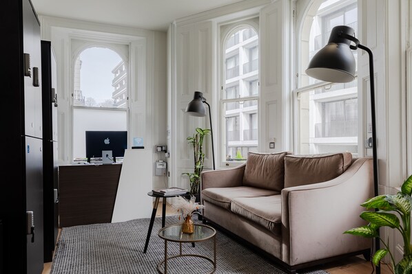 A modern living room with a beige sofa, a glass coffee table, a black floor lamp, and a view of a cityscape through large windows. - Kensington House Hotel  (London)