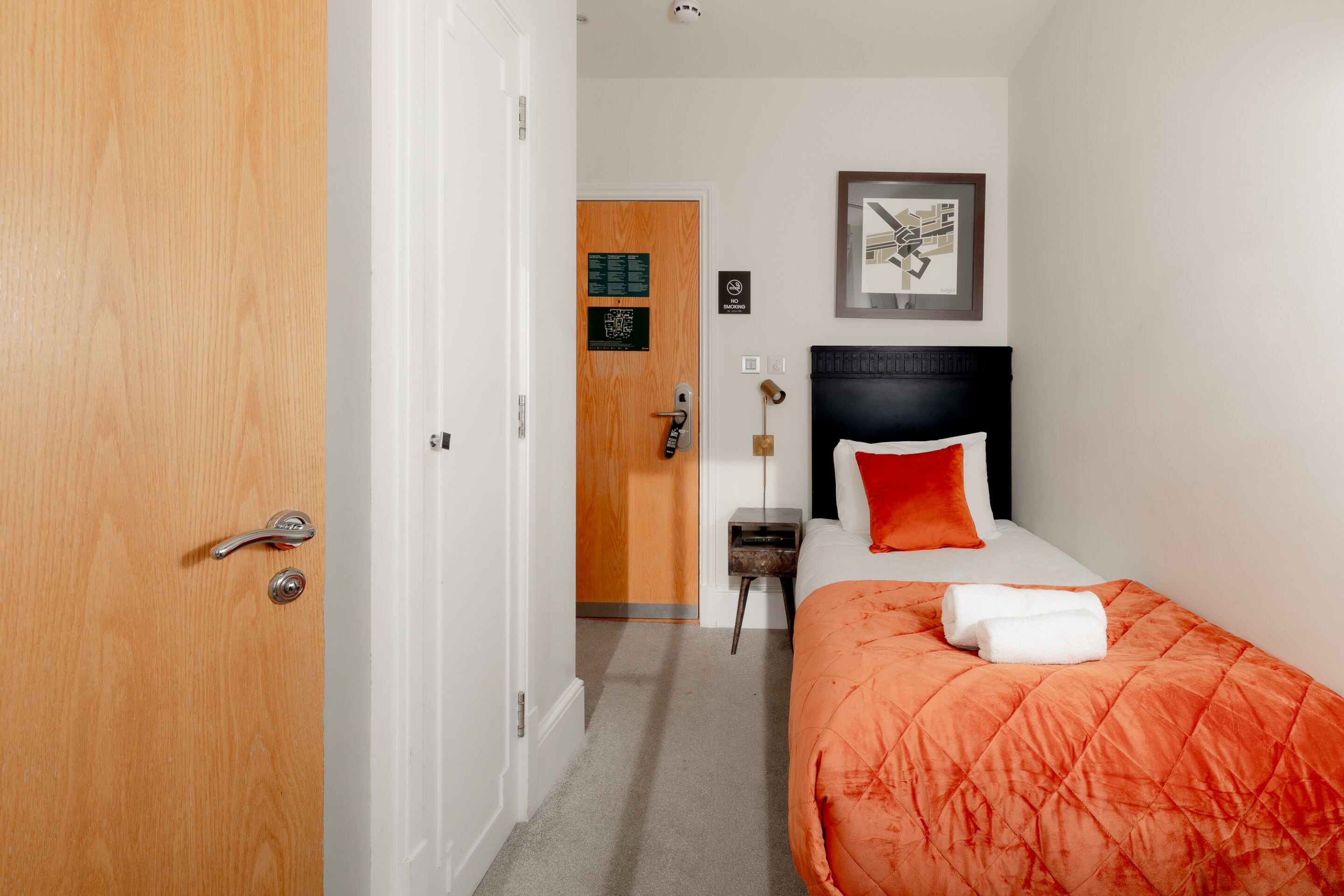 A hotel room with a wooden door, a bed with an orange quilt, a nightstand, and a framed map on the wall.
