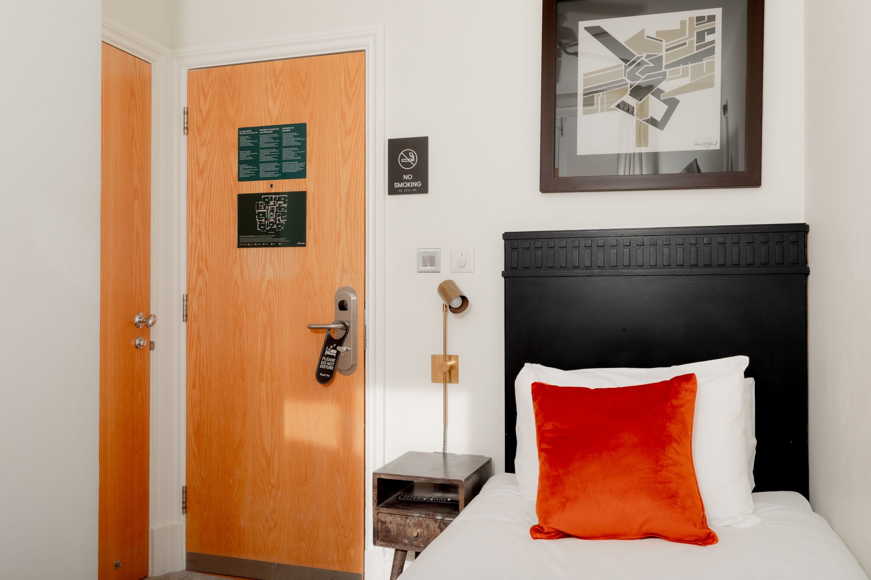 A hotel room with a wooden door, a bed with white and red pillows, a black headboard, a nightstand, a wall-mounted light, and a framed abstract artwork.