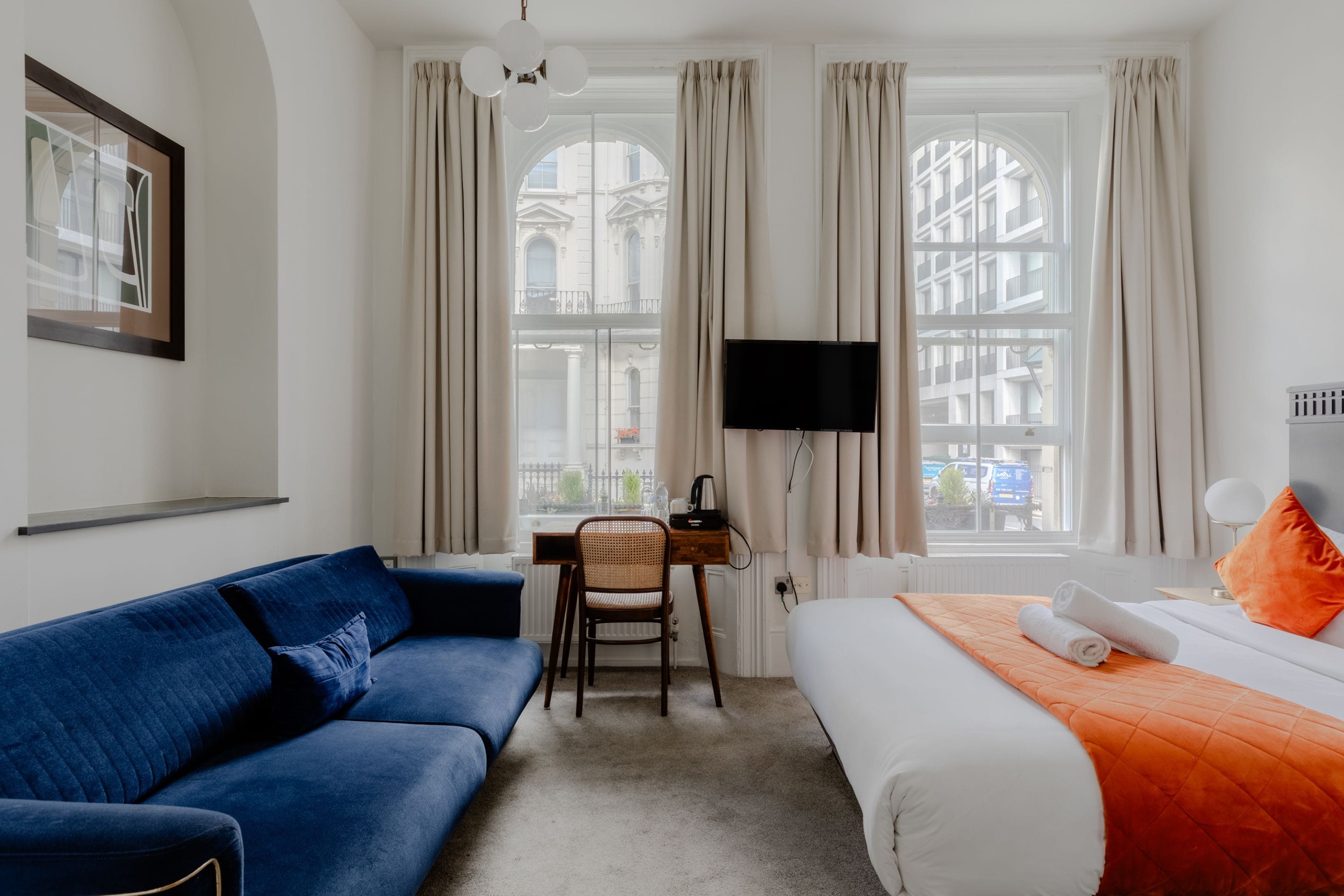A hotel room with a blue sofa, a bed with an orange bedspread, a desk with a flat-screen TV, and large windows with curtains.