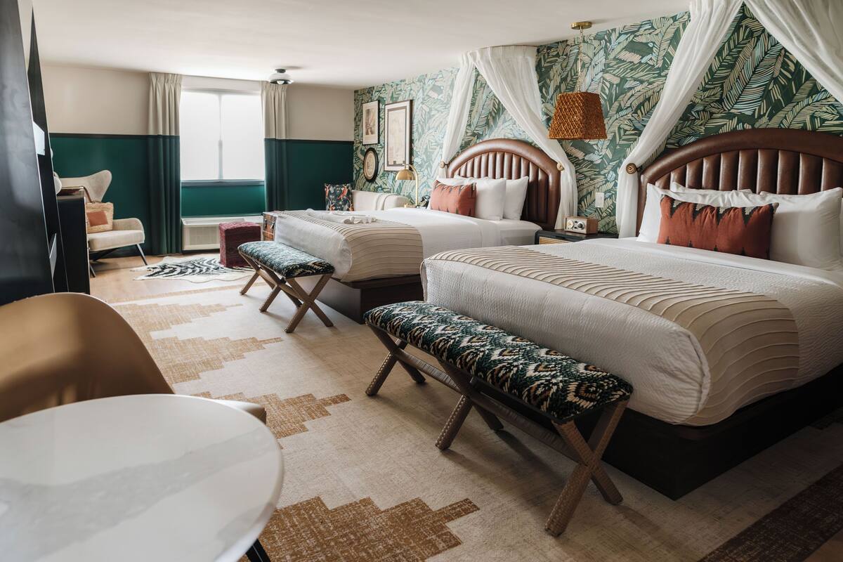 Habitación de hotel con dos camas, un banco, una silla, un escritorio y una ventana con cortinas.