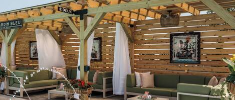 Un área de estar al aire libre con sofás verdes, cortinas blancas, una pérgola de madera y una piscina.