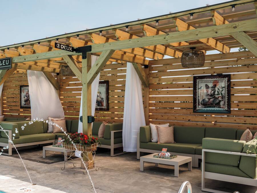Un área de estar al aire libre con sofás verdes, cortinas blancas, una pérgola de madera y una piscina.