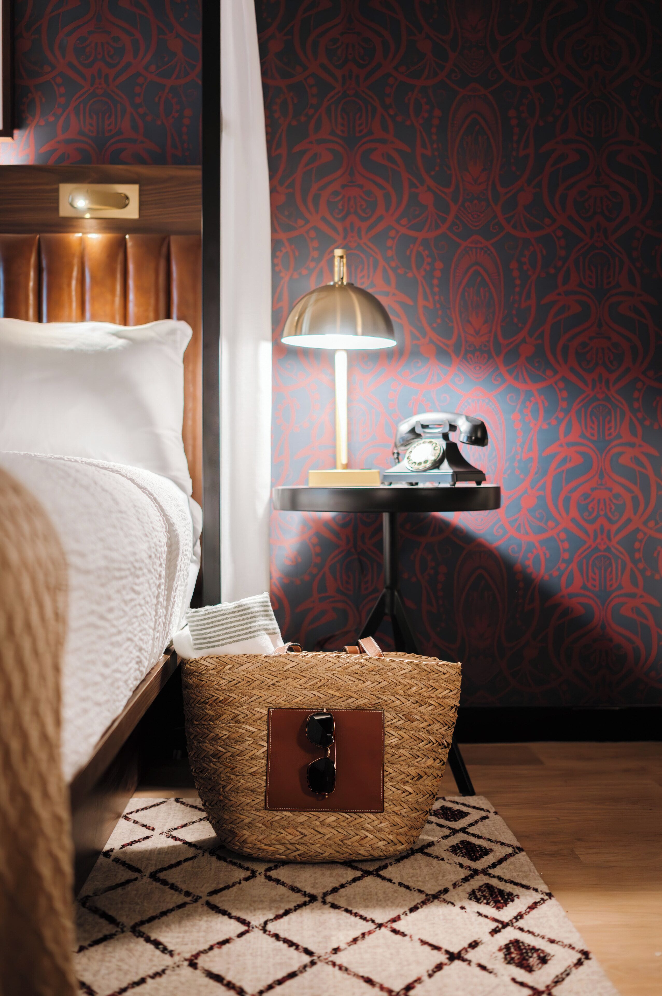 A neatly made bed with a headboard, a bedside lamp, a vintage telephone, and a woven basket with a leather pouch on a patterned rug.
