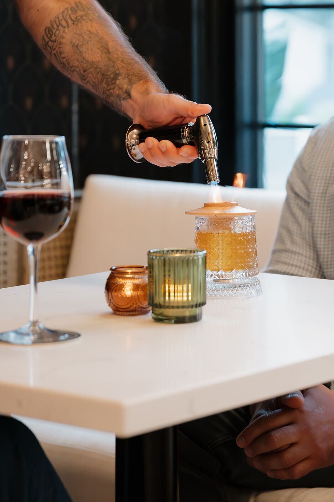 Una persona sirviendo una bebida desde un grifo en un vaso, con una vela y un recipiente decorativo sobre una mesa.