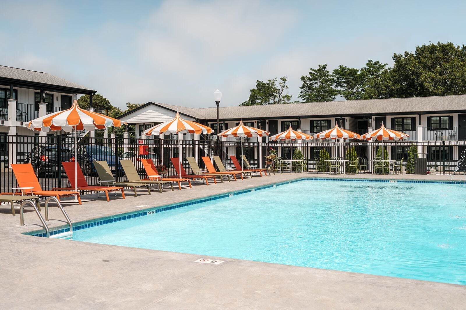 En swimmingpool med liggestole og stribede parasoller, omgivet af et hegn og bygninger.