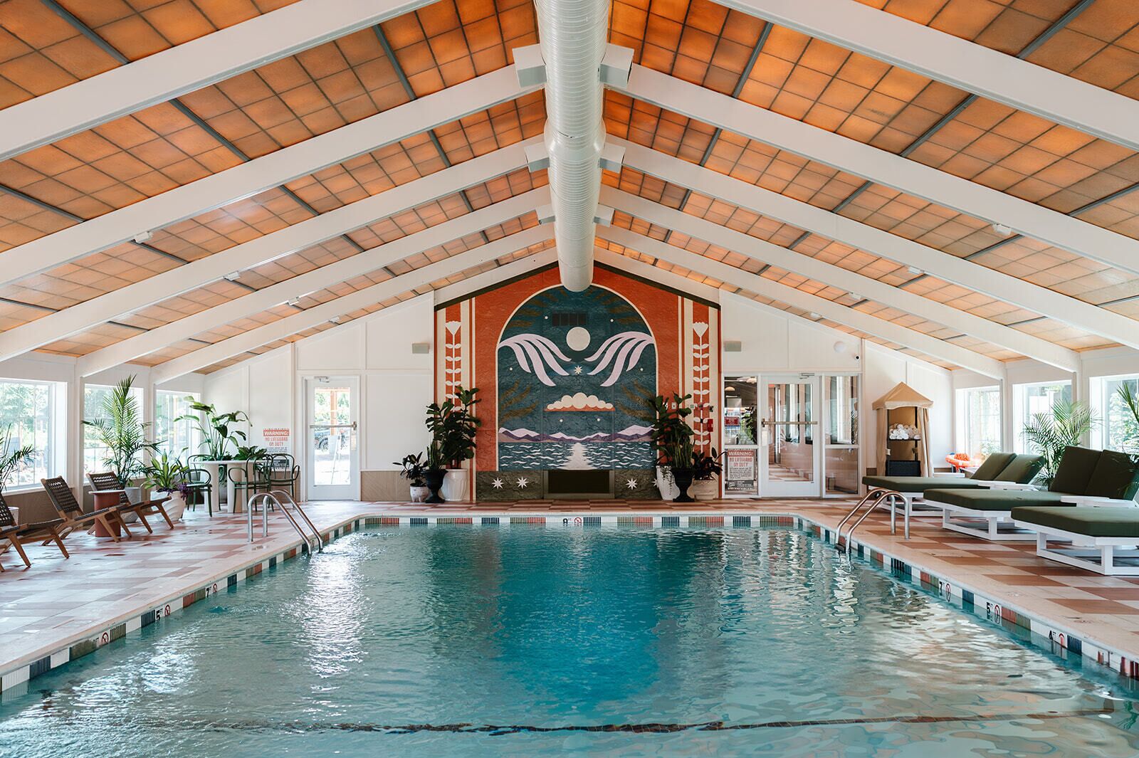 An indoor swimming pool with a high ceiling, lounge chairs, and potted plants.