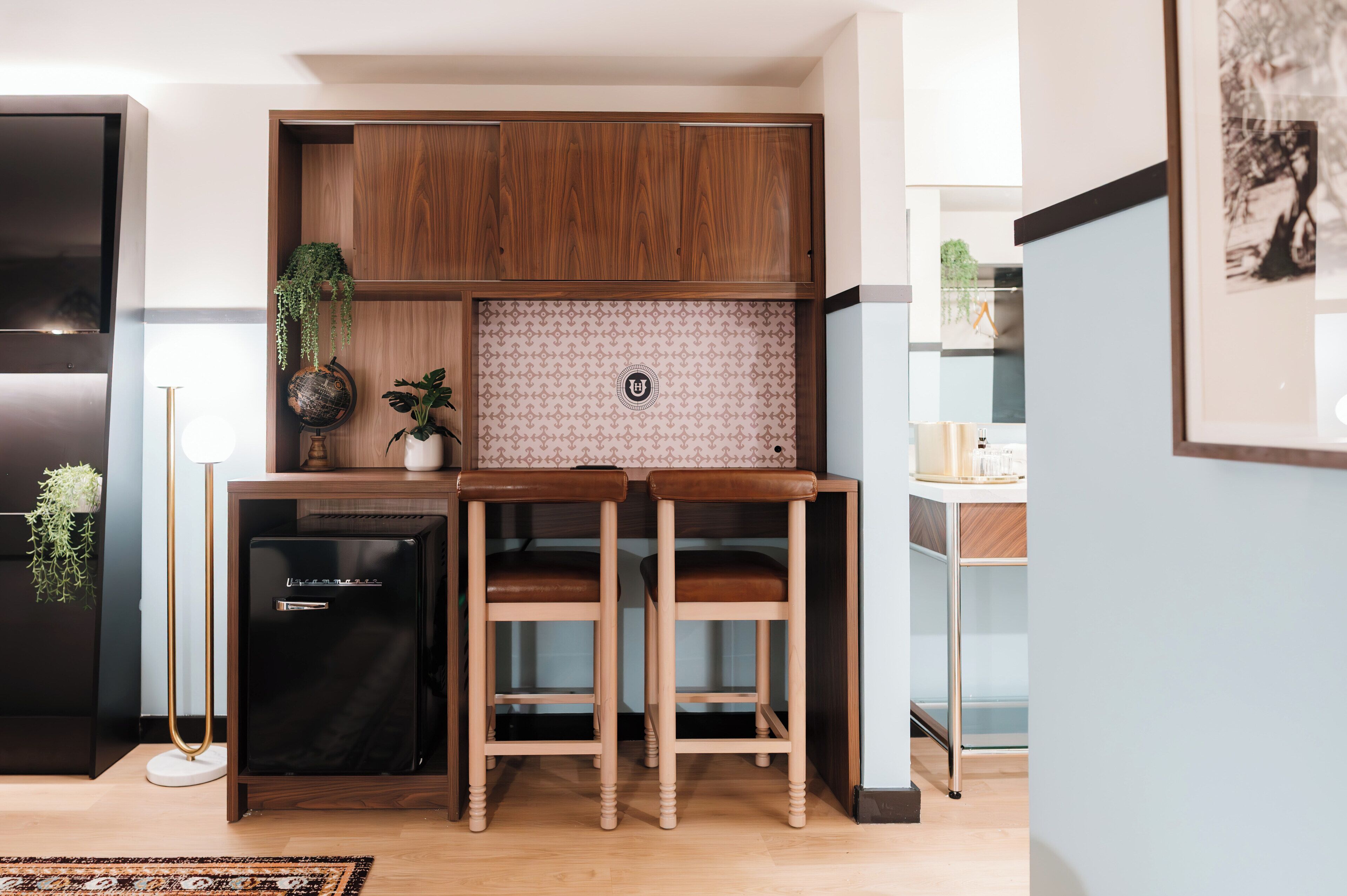 A compact kitchen with wooden cabinets, a small refrigerator, two bar stools, and a potted plant.