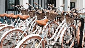 A row of white bicycles with brown seats and wicker baskets, parked in front of a white building with black railings and large windows. - Uncommoner Hotel by Reverie Boutique Collection (West Yarmouth)