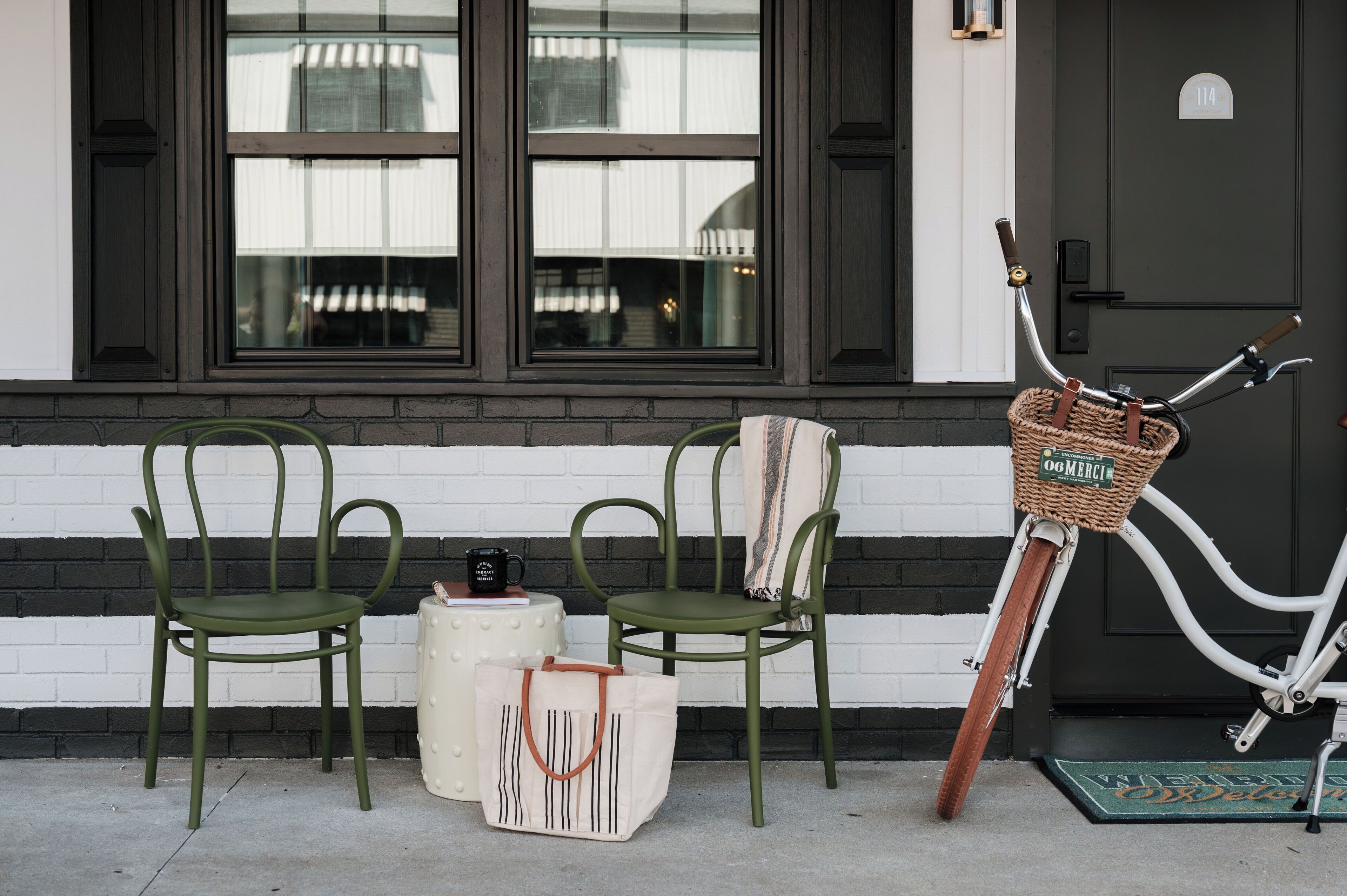 Two green chairs, a white table with a black mug, a white tote bag, and a wicker bike basket with a handle.