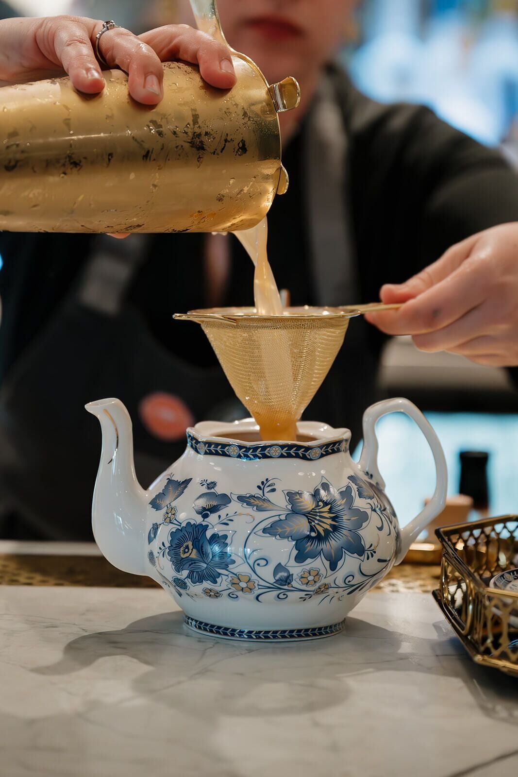 Iemand giet een vloeistof uit een kan in een theepot met een bloemenmotief.