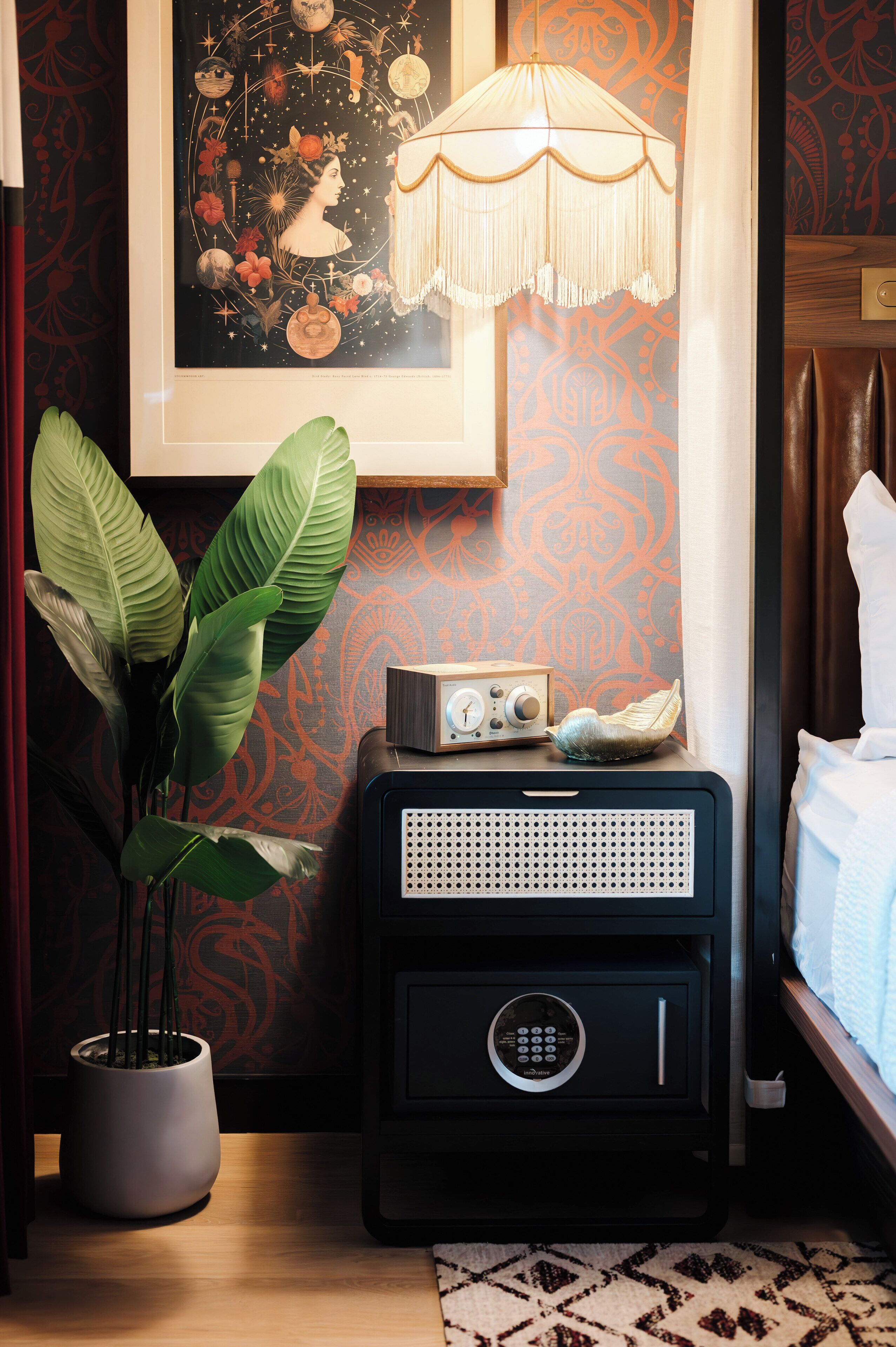 Une pièce avec une plante en pot, une radio ancienne, un tableau encadré et un lit recouvert d’une couette blanche.