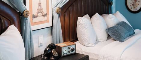 A bedroom with a wooden headboard, white bedding, a telephone, a radio, and a framed picture of the Eiffel Tower.
