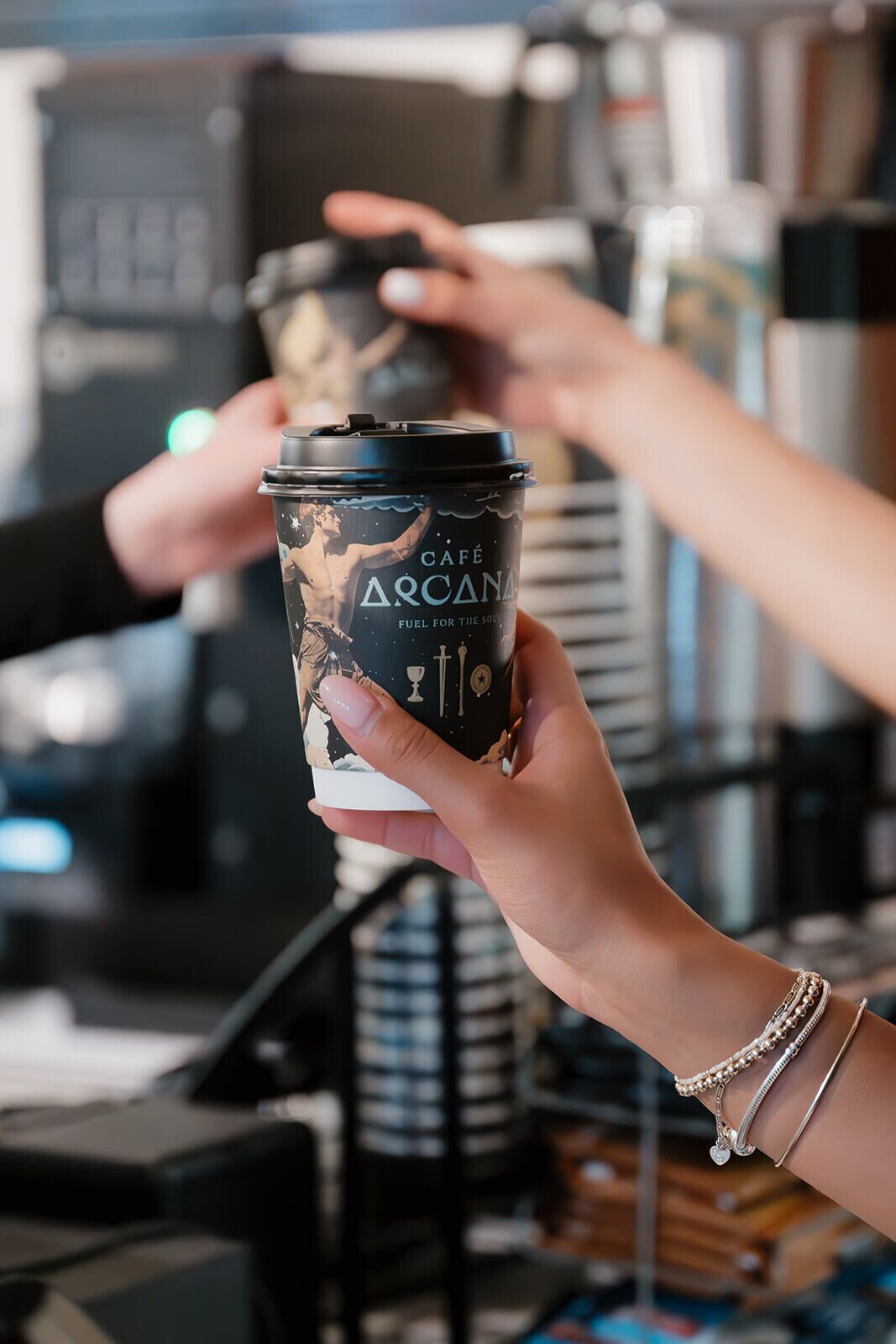 Eine Person hält eine Kaffeetasse, auf der das Label 'CAFÉ ARCANA FUEL FOR THE SOUL' steht.