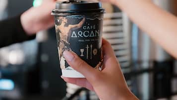 A person holding a coffee cup with a label that reads 'CAFÉ ARCANA FUEL FOR THE SOUL.'