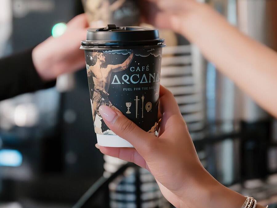 A person holding a coffee cup with a label that reads 'CAFÉ ARCANA FUEL FOR THE SOUL.'