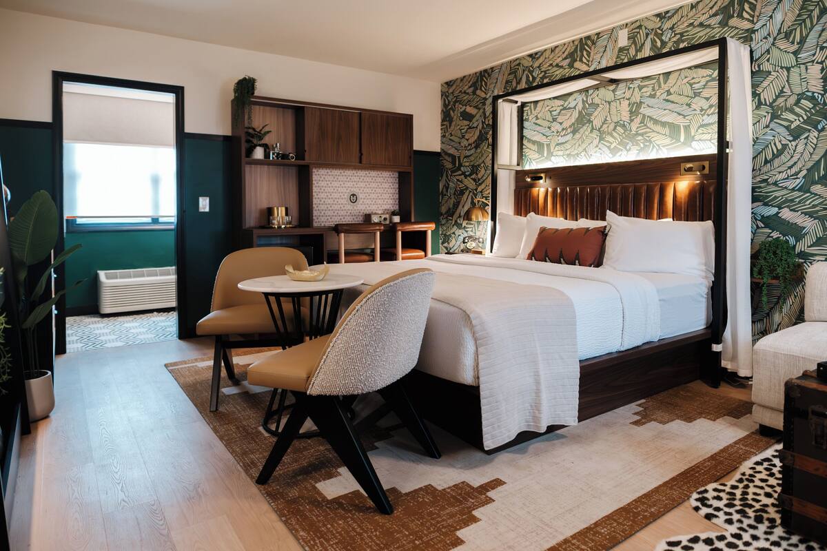 Habitación de hotel con una cama grande, un escritorio, una silla y una mesita. Se ve una ventana con persianas, una planta y un papel tapiz estampado.