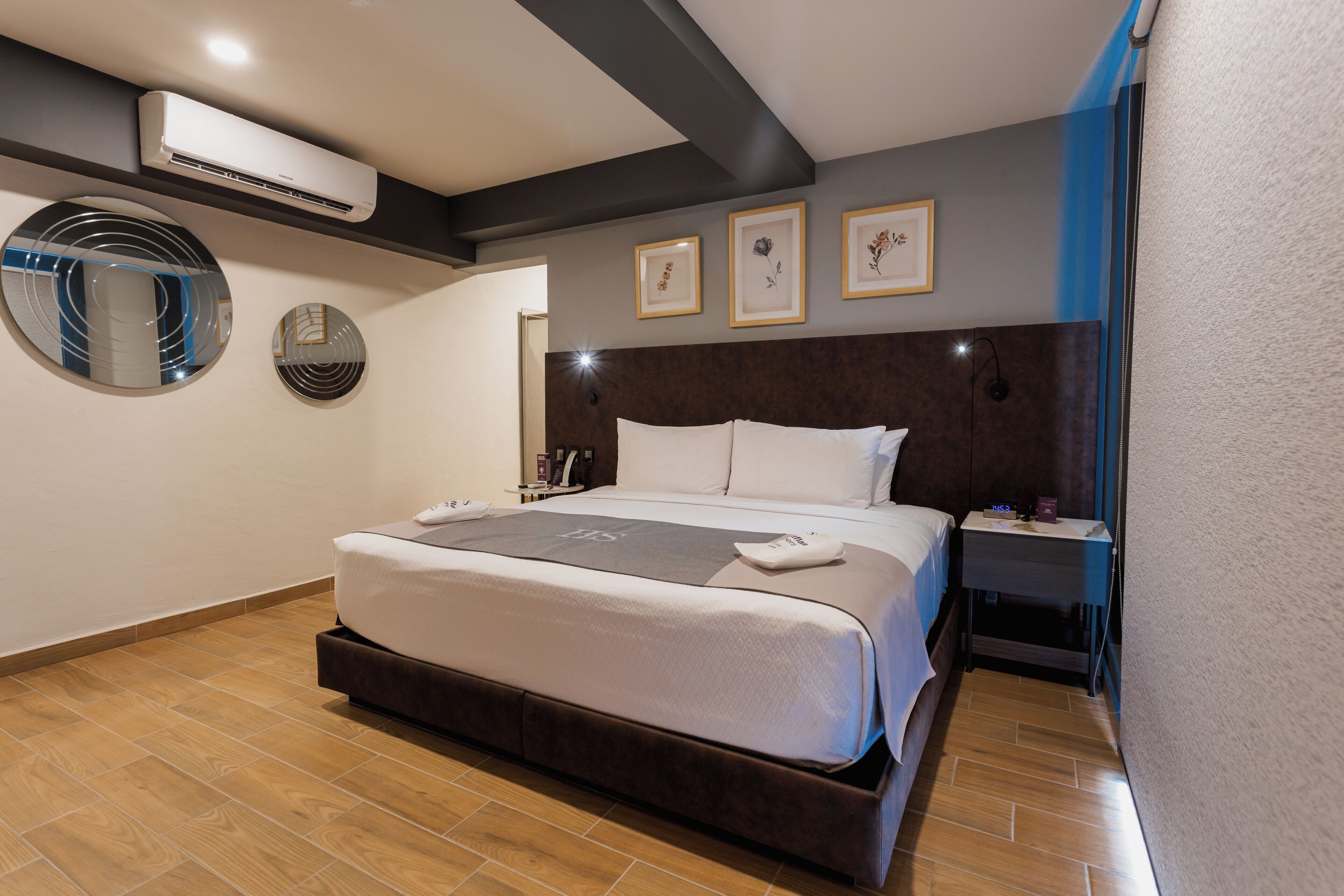 A modern hotel room with a large bed, a bedside table, a wall-mounted air conditioner, and framed artwork on the wall.