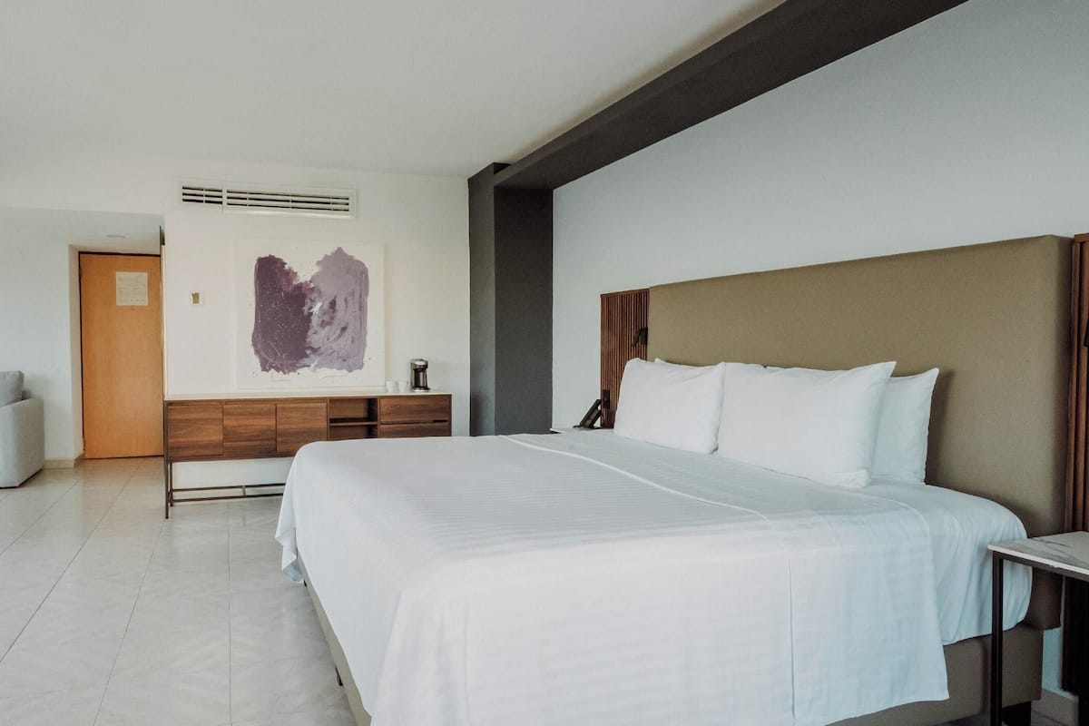 A modern hotel room with a large bed, a desk, a sofa, and a wall-mounted artwork.
