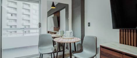 Una sala de estar moderna con un ventanal que da a un balcón, una mesa redonda de madera con dos sillas, un televisor de pantalla plana montado en la pared y un mueble de madera.