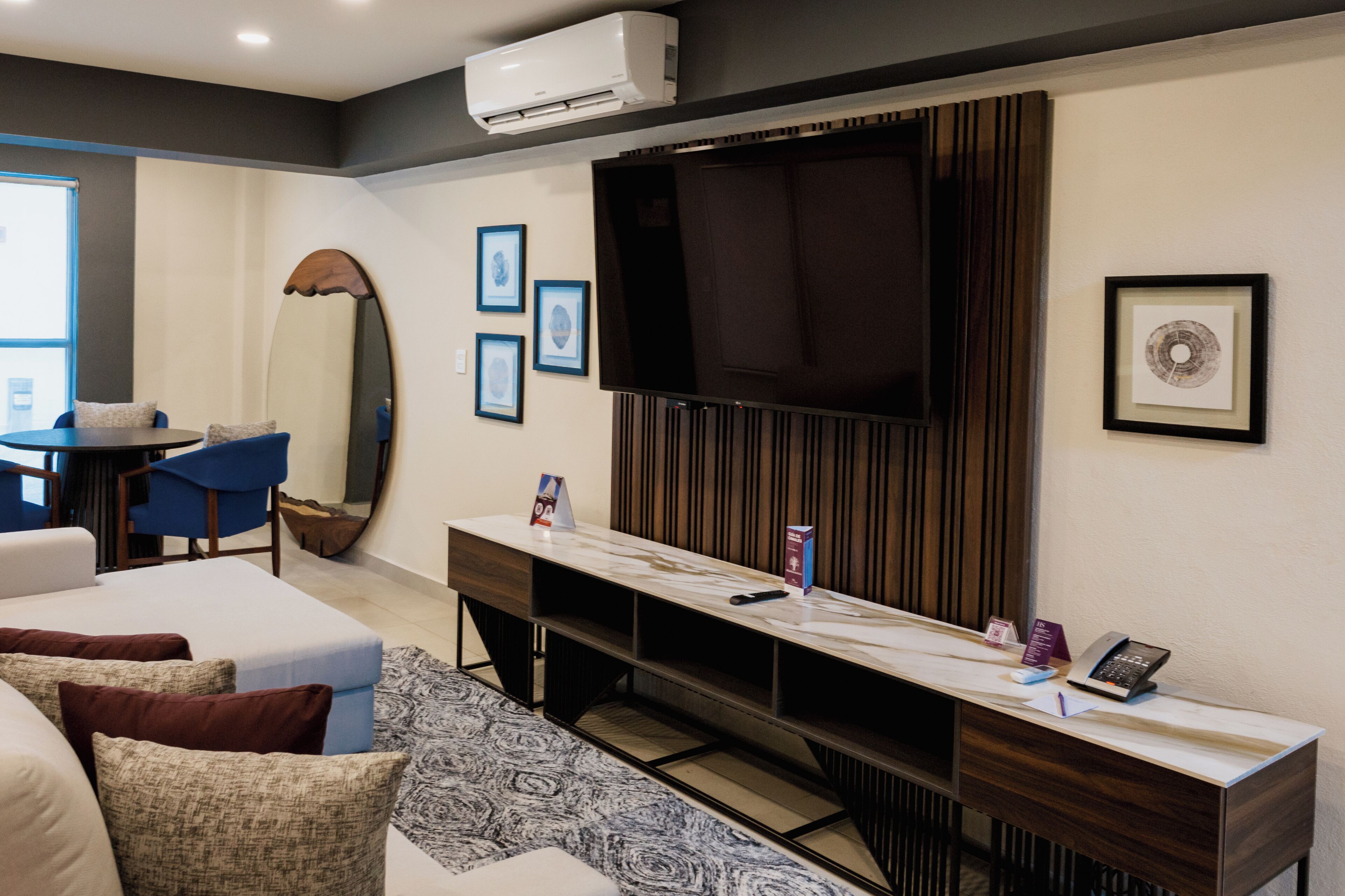 A modern hotel room with a sofa, a desk with a flat-screen TV, a large mirror, and framed pictures on the wall.
