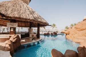 Zona de piscina con un bar de techo de paja llamado 'COCO'S BAR', rodeado de rocas y palmeras.