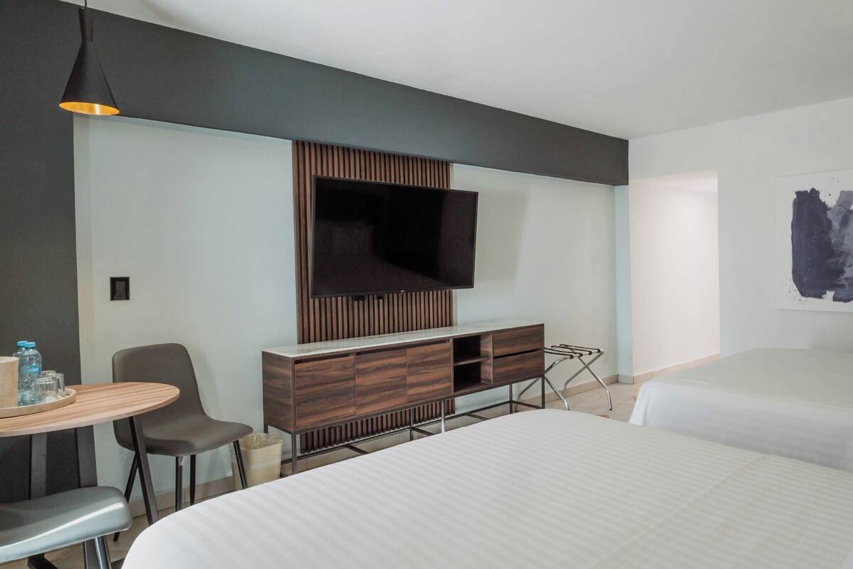 A hotel room with a bed, a TV mounted on a wooden panel, a desk with a chair, and a painting on the wall.