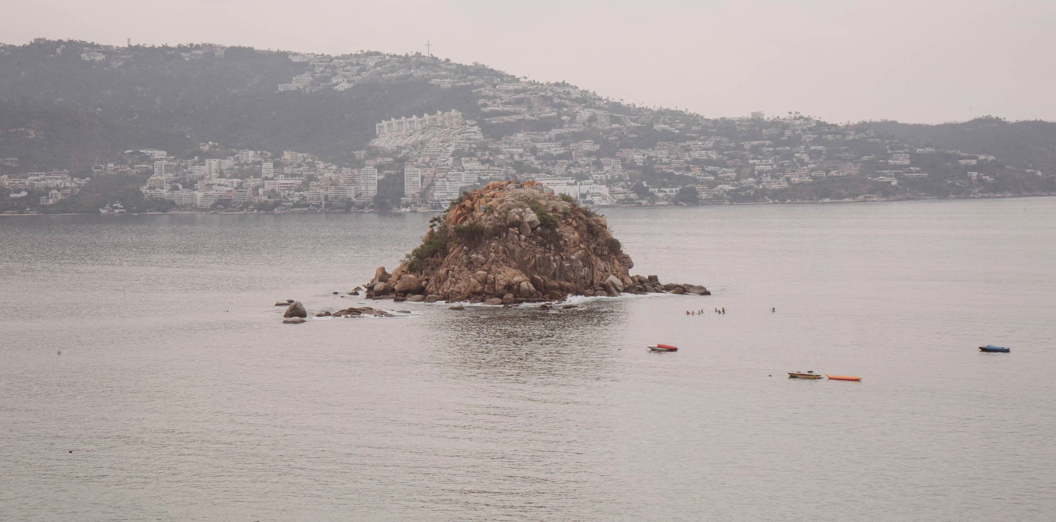 A rocky island in a body of water with buildings in the background.