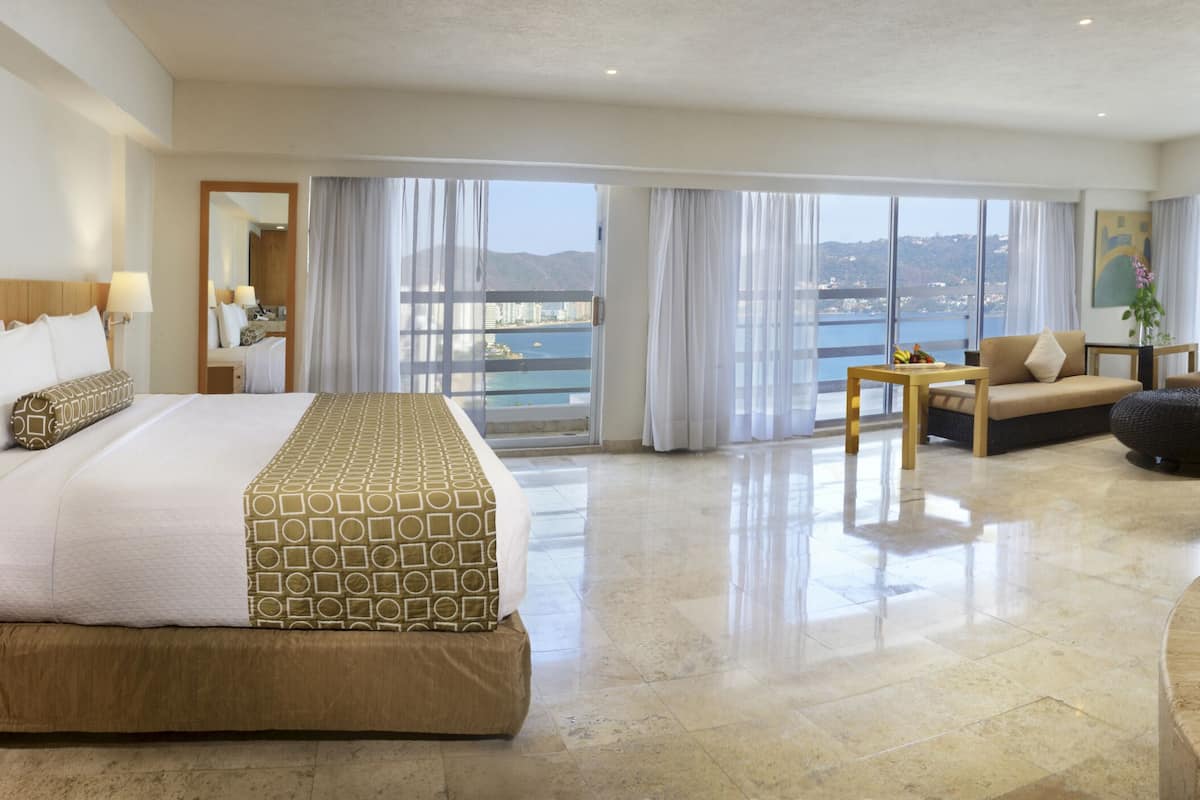 A modern hotel room with a large bed, a sitting area with a sofa and armchairs, a separate bathroom with a bathtub, and a balcony with a view of the city.