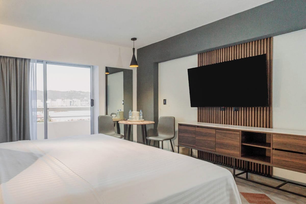 A hotel room with a large bed, a desk, a chair, a TV, and a view of the city through the window.